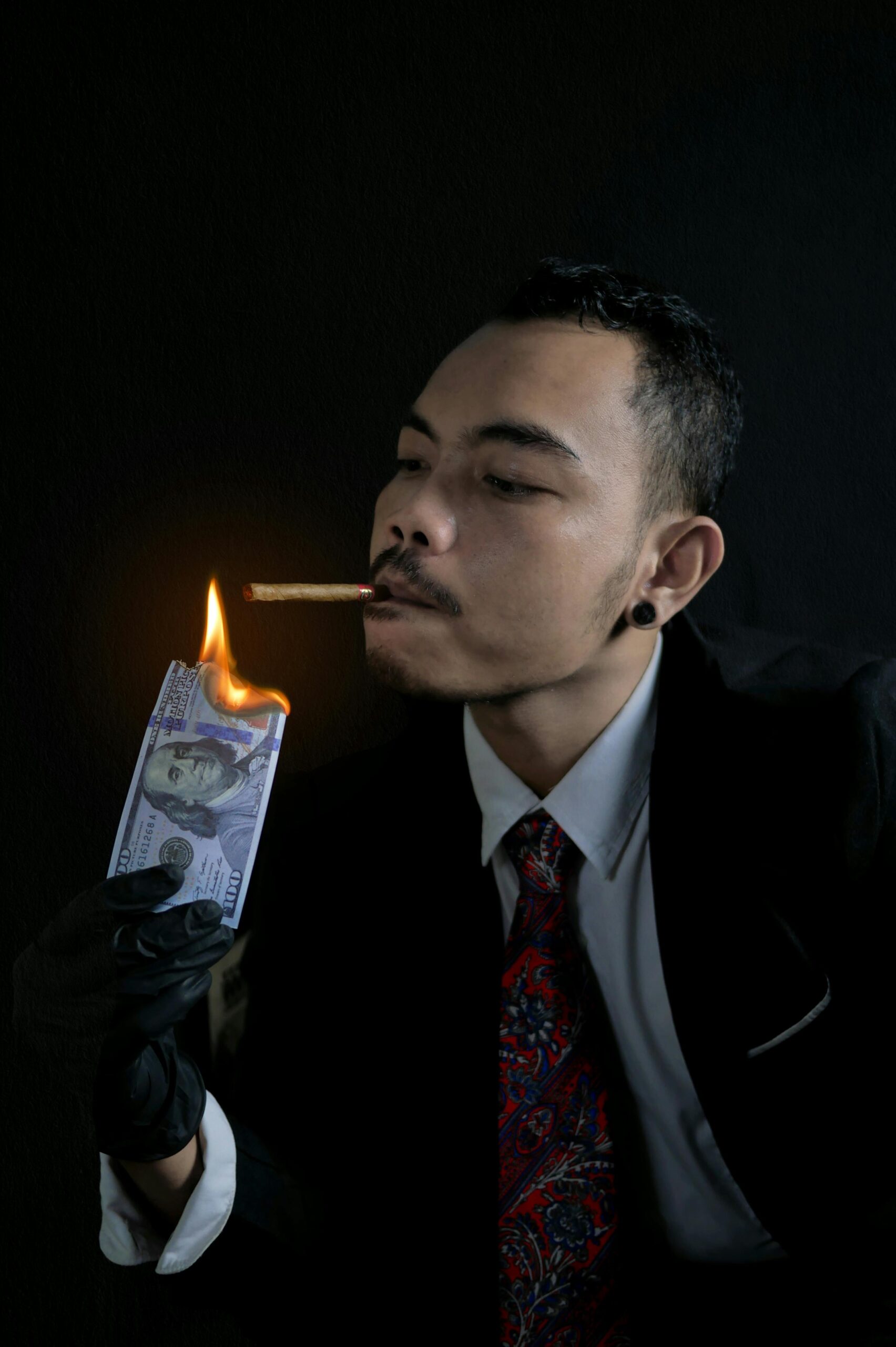 Portrait of a man using a burning banknote to light a cigarette, wearing business attire and gloves.
