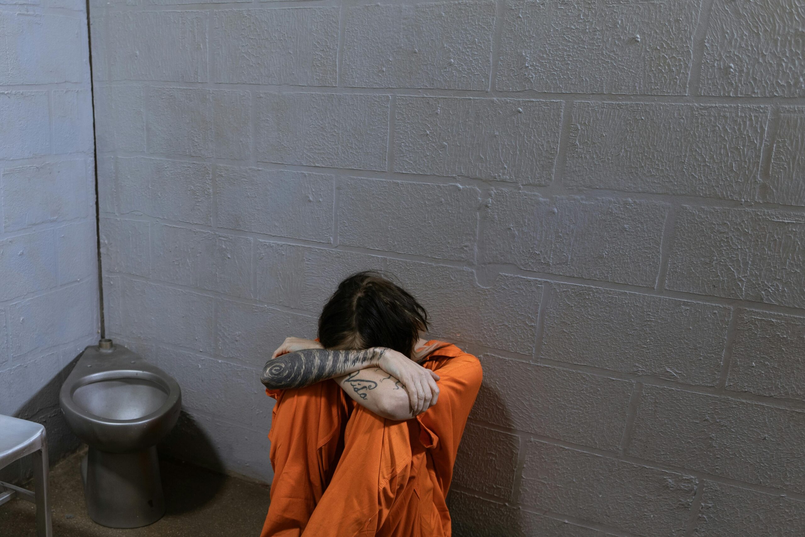 A solitary prisoner in orange uniform sits on the floor of a sparse jail cell.